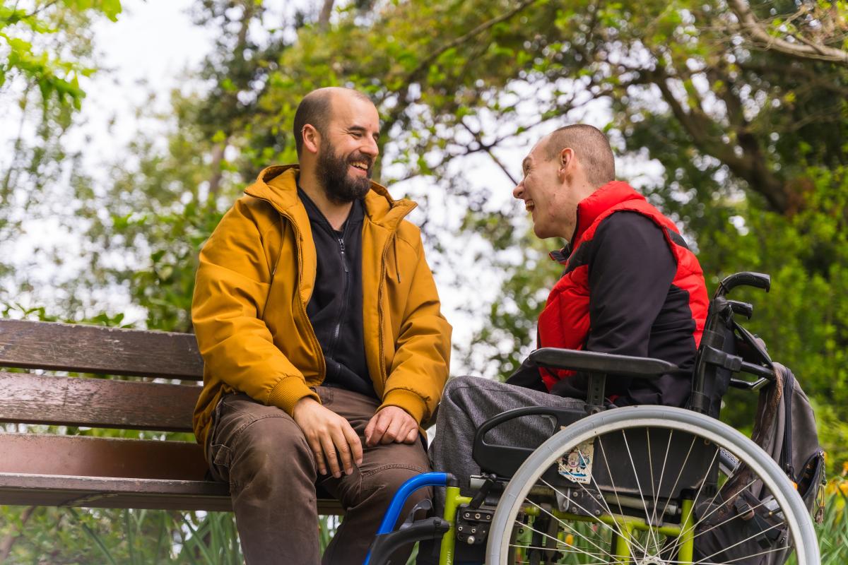 Zwei Männer im Park, einer auf Band sitzend, der andere davor im Rollstuhl