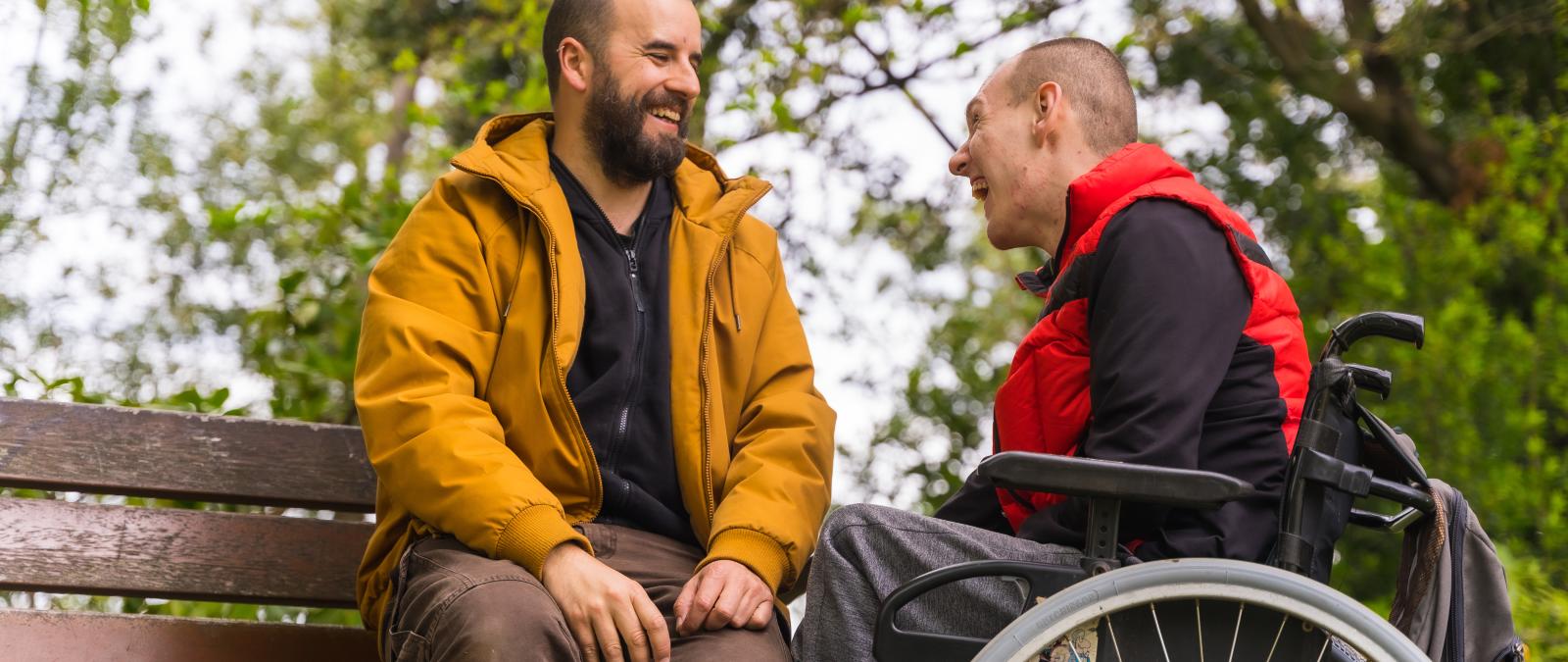 Zwei Männer im Park, einer auf Band sitzend, der andere davor im Rollstuhl